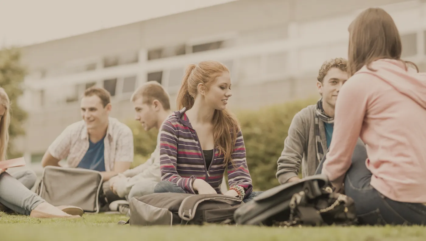 A group of young students sitting on the grass outside a campus building, talking together